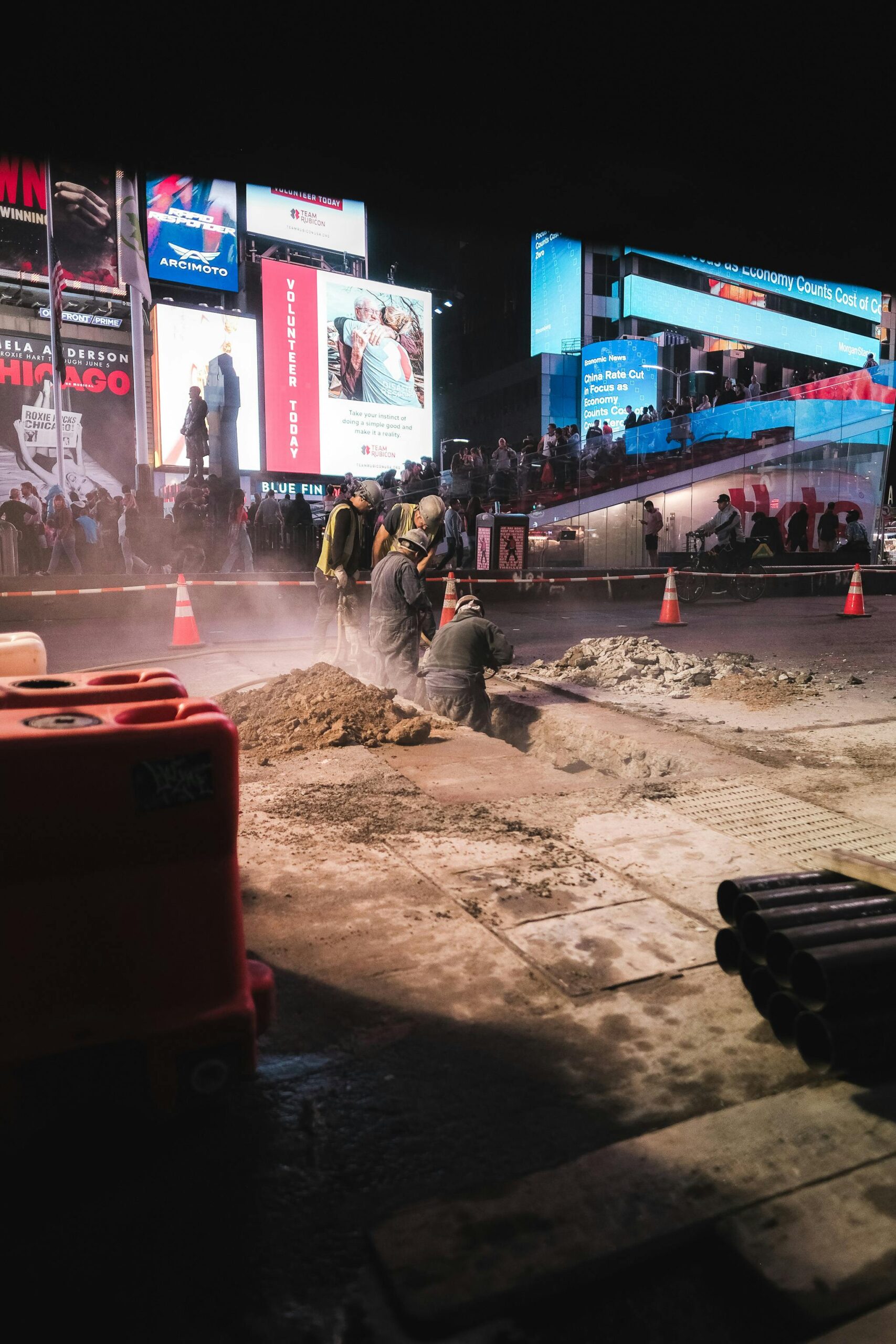 Workers engaging in nighttime road construction at a vibrant city intersection with lighted billboards.